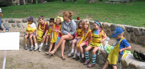 bambini con casacche gialle per identificarli seduti su un muretto, accanto a un prato, controllati da un'accompagnatrice