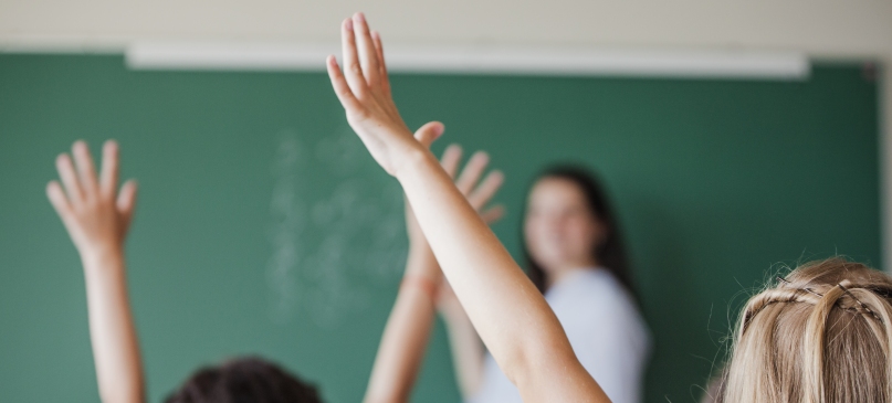 bambini in aula con le mani alzate e maestra alla lavagna sullo sfondo