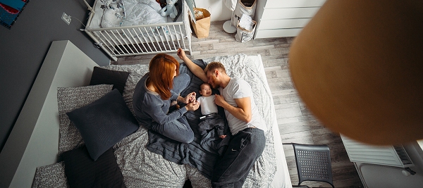 coppia di giovani genitori seduti su un letto, tra di loro un bambino di pochi mesi