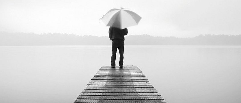 uomo su un porticciolo, di spalle, sotto ad un ombrello, solo, in una foto in biancoe nero