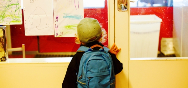 bimbo di 3 o 4 anni, visto di schiena, che si appoggia alla porta della scuola nella quale sta per entrare