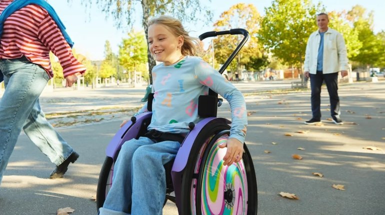 bambina in strada sorridente sulla sua sedia a rotelle. vicino a lei si intravedono due persone