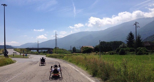 due ragazzi su handbike, sullo sfondo cielo azzurro e montagne 