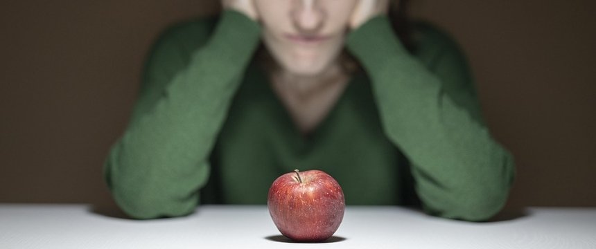 sagoma di una persona seduta, con il viso appoggiato sulle mani, e davanti a lei una mela su un tavolo
