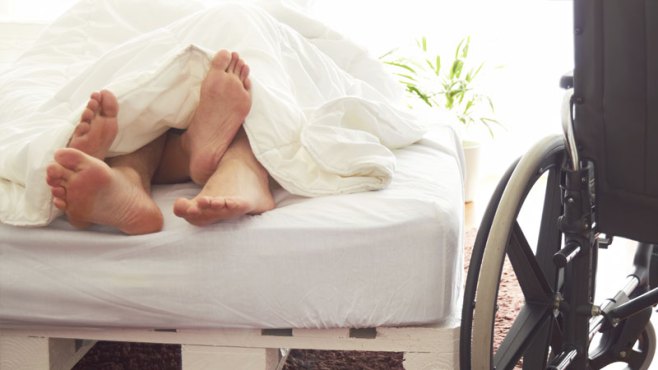 piedi di uomo e donna che escono dalle coperte alla fine del letto, accanto al quale è collocata una sedia a rotelle