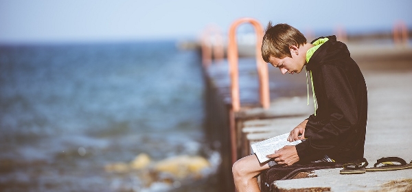 ragazzo seduto su un molo che sta leggendo un libro con la schiena ricurva
