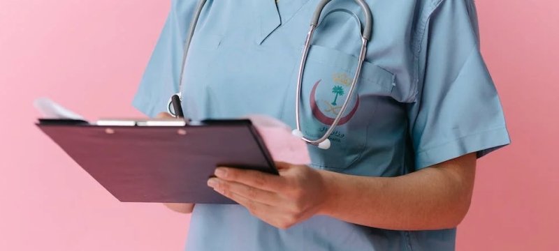 dettaglio delle mani di un infermiere che scrive su una cartella, indossando un camice azzurro e lo stetoscopio sul collo