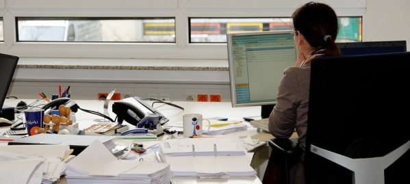 donna vista di schiena al lavoro, seduta ad una scrivania piena di documenti sparsi ovunque