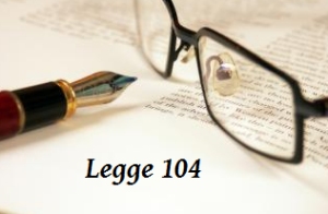 dettaglio di un paio di occhiali appoggiati su un foglio vicino a una penna e un foglio, e la scritta legge 104